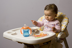 Baby im Hochstuhl sitzend mit Trinkbecher, greift nach Fingerfood.